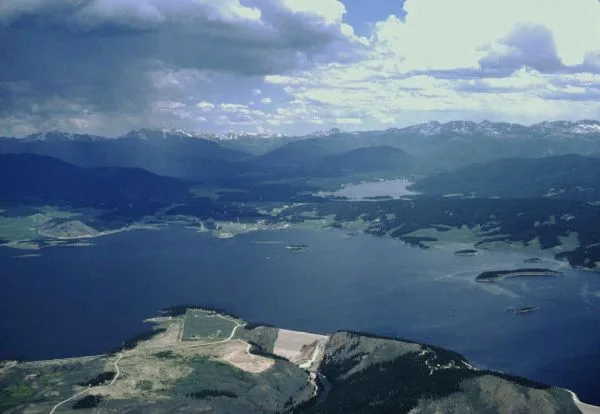 Wide aerial view over Grand Lake with dark blue water filling the frame, a small town and shoreline at the lower edge, and mountain ridges rising in the distance. Broken clouds and bright morning light create a hazy, atmospheric scene with a hint of reflection on the lake surface.