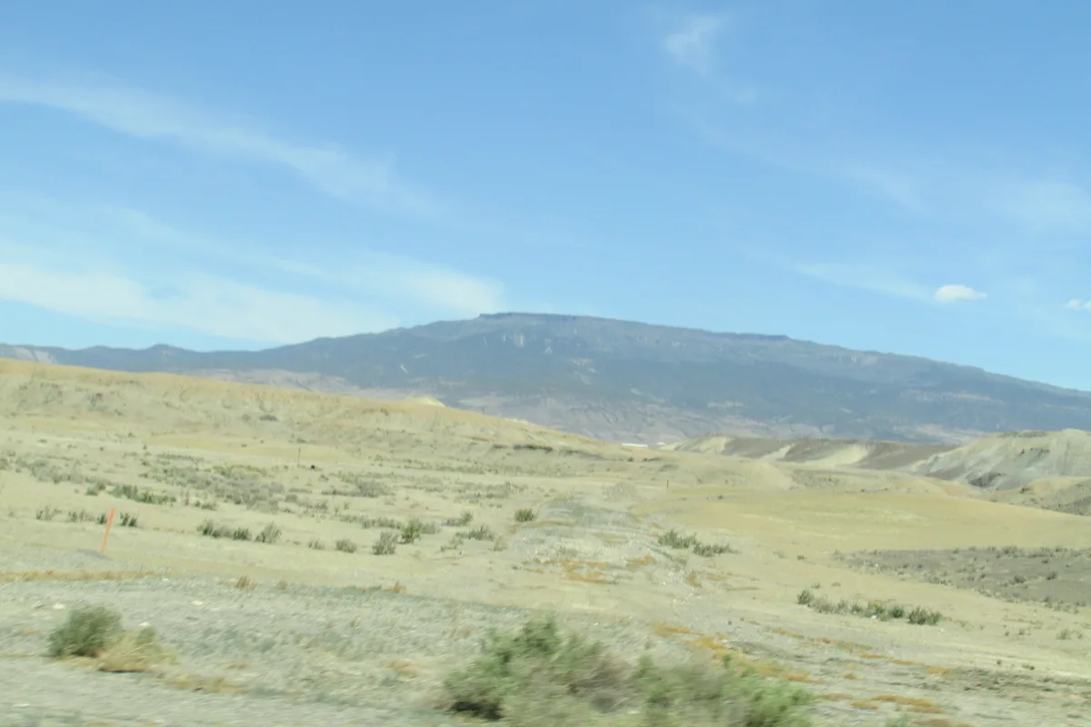 Wide view across the Grand Mesa’s pale, open plateau under a clear blue sky, with low scrub and rolling hills leading to a dark, flat-topped ridgeline in the distance. The scene has the soft light of late day, with a muted golden tint across the landscape.