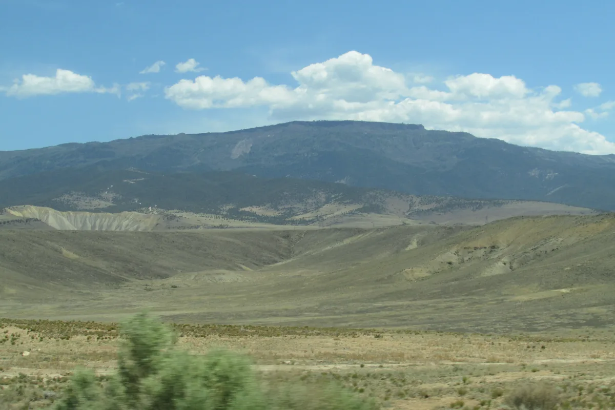 Wide view across Grand Mesa’s broad, treeless plateau with rolling tan and green ridges in the foreground and a dark, flat-topped mountain stretching across the horizon. Puffy white clouds float in a bright blue sky, giving the scene a clear midday feel.