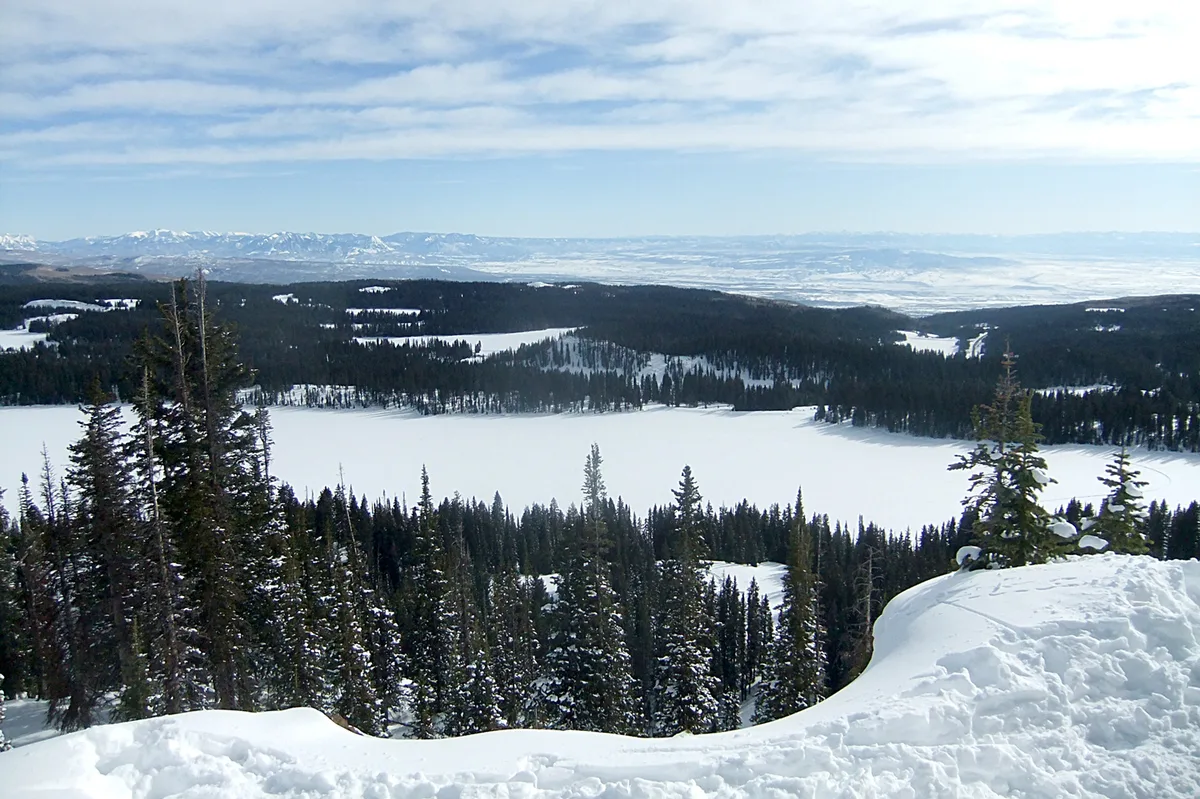 Snow-covered Grand Mesa stretches across the frame, with frozen alpine lakes and dark evergreen forests layered below a pale blue sky. Distant mountain ridges line the horizon, while a rounded snowbank in the foreground frames the overlook.