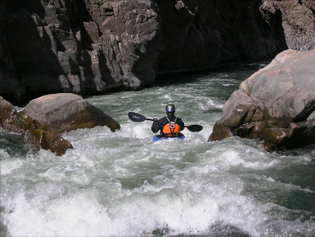 A kayaker in an orange life vest paddles through frothy whitewater between huge dark canyon walls and rounded boulders in the Gunnison Gorge. Morning light catches the churning river while the rock faces remain mostly in deep shadow.