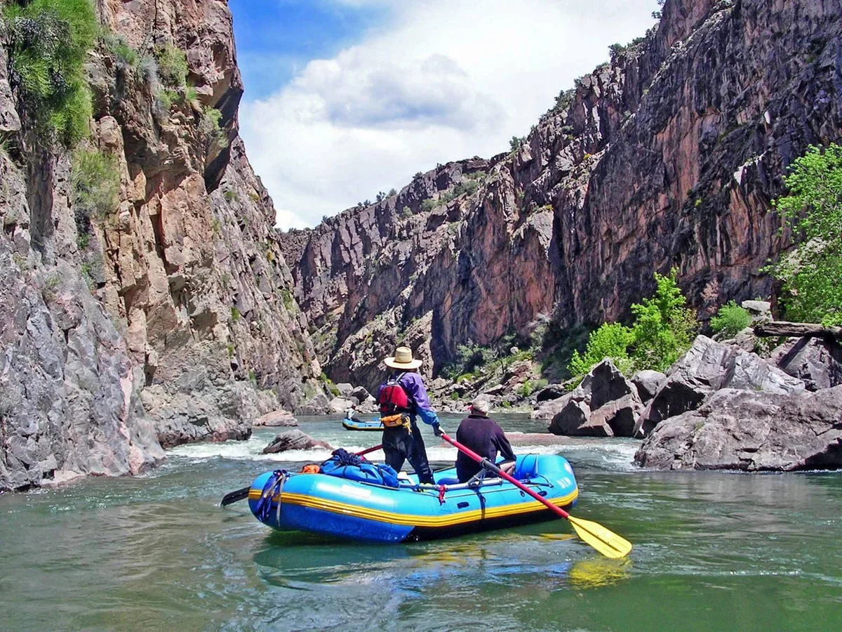 Two people float a blue inflatable raft through a narrow canyon on calm green water, surrounded by steep dark rock walls and scattered boulders. Bright morning light and a partly cloudy sky illuminate the rugged gorge and a few green shrubs clinging to the rock.