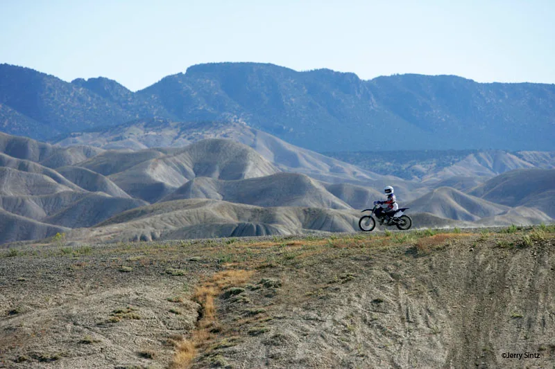 A motorcyclist rides along the narrow top of a dry, rocky ridge above a steep canyon drop, with layered gray badlands and blue mountains stretching across the background. The low morning light highlights the eroded slopes and the remote, open feel of the Gunnison Gorge wilderness.