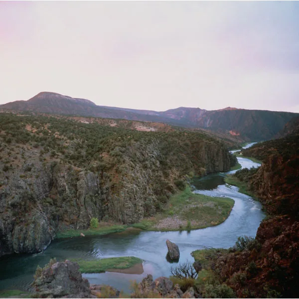 A winding river cuts through a steep, rocky canyon lined with dark cliffs and sparse scrub, with low mountains stretching across the horizon under a pale morning sky. The calm water curves around gravel bars and a lone boulder, emphasizing the remote, rugged wilderness below.