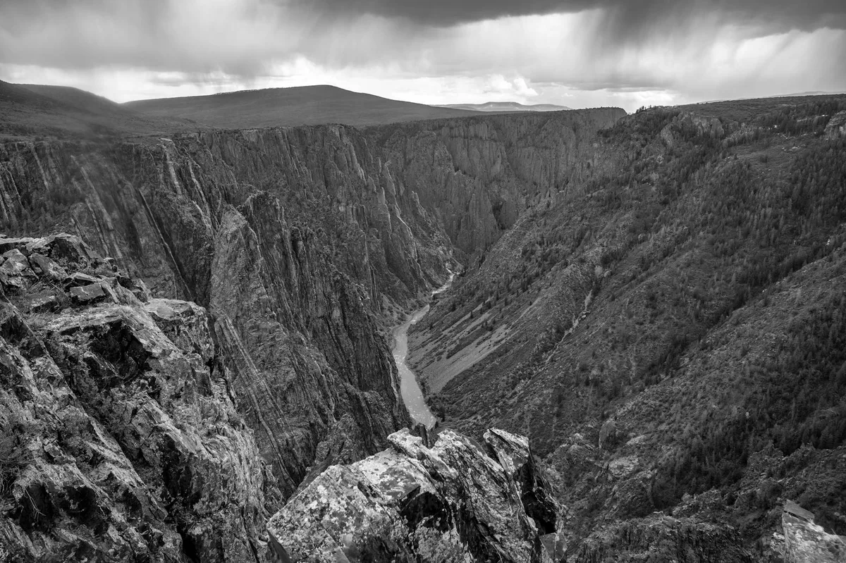 A wide black-and-white view looks down into a steep, remote river gorge with sheer dark rock walls and a narrow ribbon of river winding far below. Jagged foreground rocks frame the scene, while heavy clouds and soft morning light emphasize the canyon’s depth and rugged texture.
