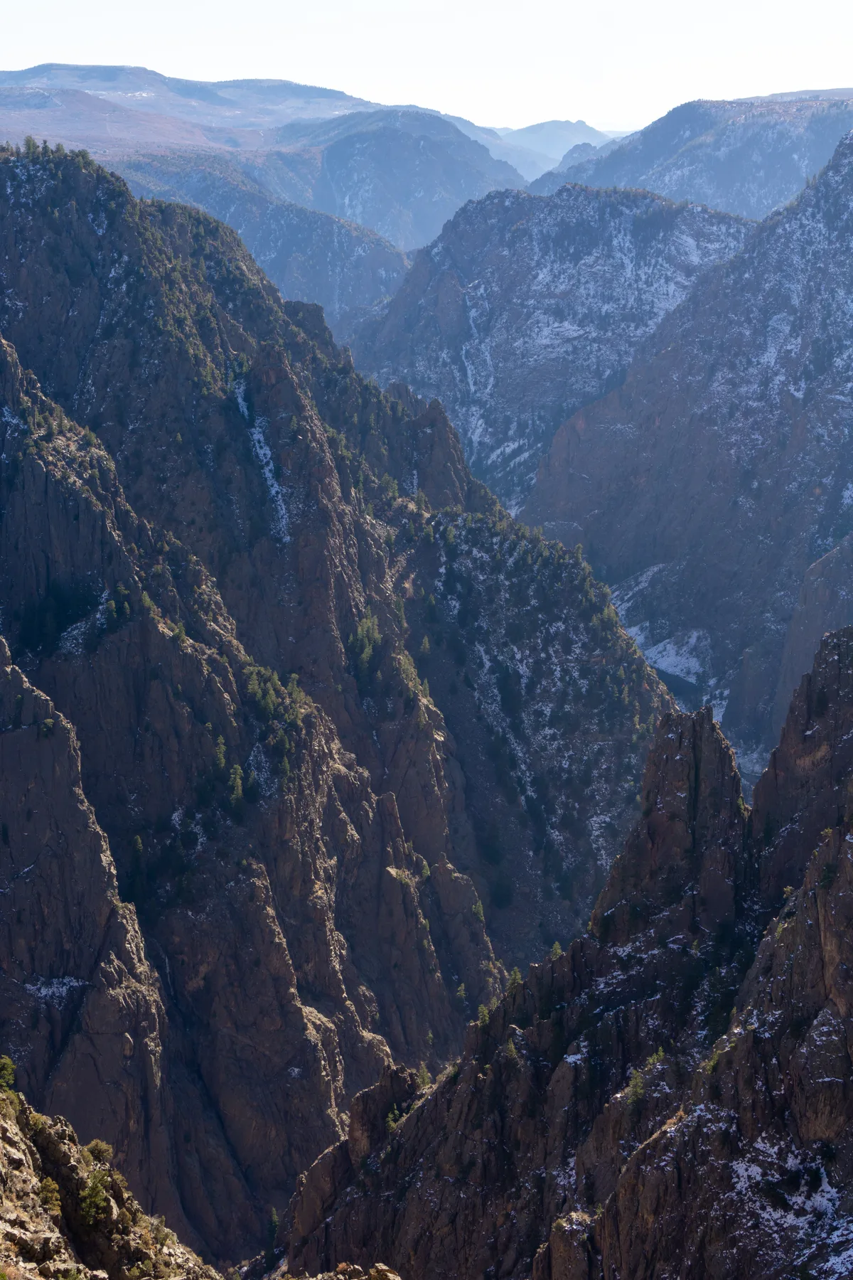 Steep, jagged canyon walls plunge into a narrow gorge, with patches of snow clinging to dark rock and scattered evergreens on the ridges. Pale winter light and layered blue mountains in the distance emphasize the canyon’s immense depth.