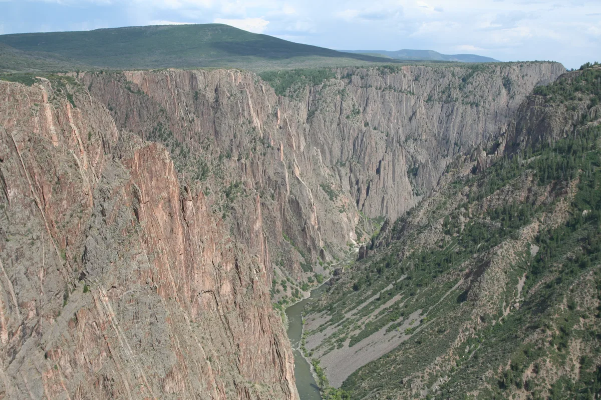 Sheer rust-and-gray canyon walls plunge toward a narrow green ribbon of the Gunnison River, with steep, shadowed slopes and sparse pines clinging to the rocky rim. Pale blue sky and distant mesas stretch above the deep gorge, emphasizing the canyon’s scale and depth.