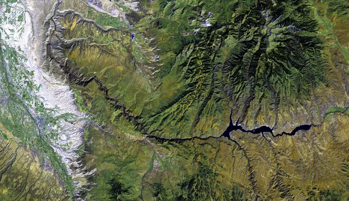A satellite-style map view shows the Black Canyon area as a deep, dark, narrow gorge cutting across rugged green and tan terrain, with steep ridges and river channels radiating outward. Pale snow-covered mountains lie to the left, contrasting with the sharply carved canyon walls and reservoir-like waterway running through the center.