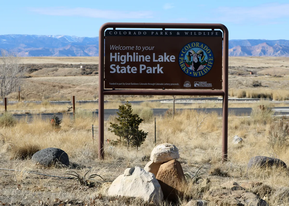 A large “Welcome to your Highline Lake State Park” sign stands in a dry, grassy high-desert foreground, with low hills and the blue-gray Book Cliffs stretching across the background. Pale winter light and a clear sky give the scene a crisp, open feel.