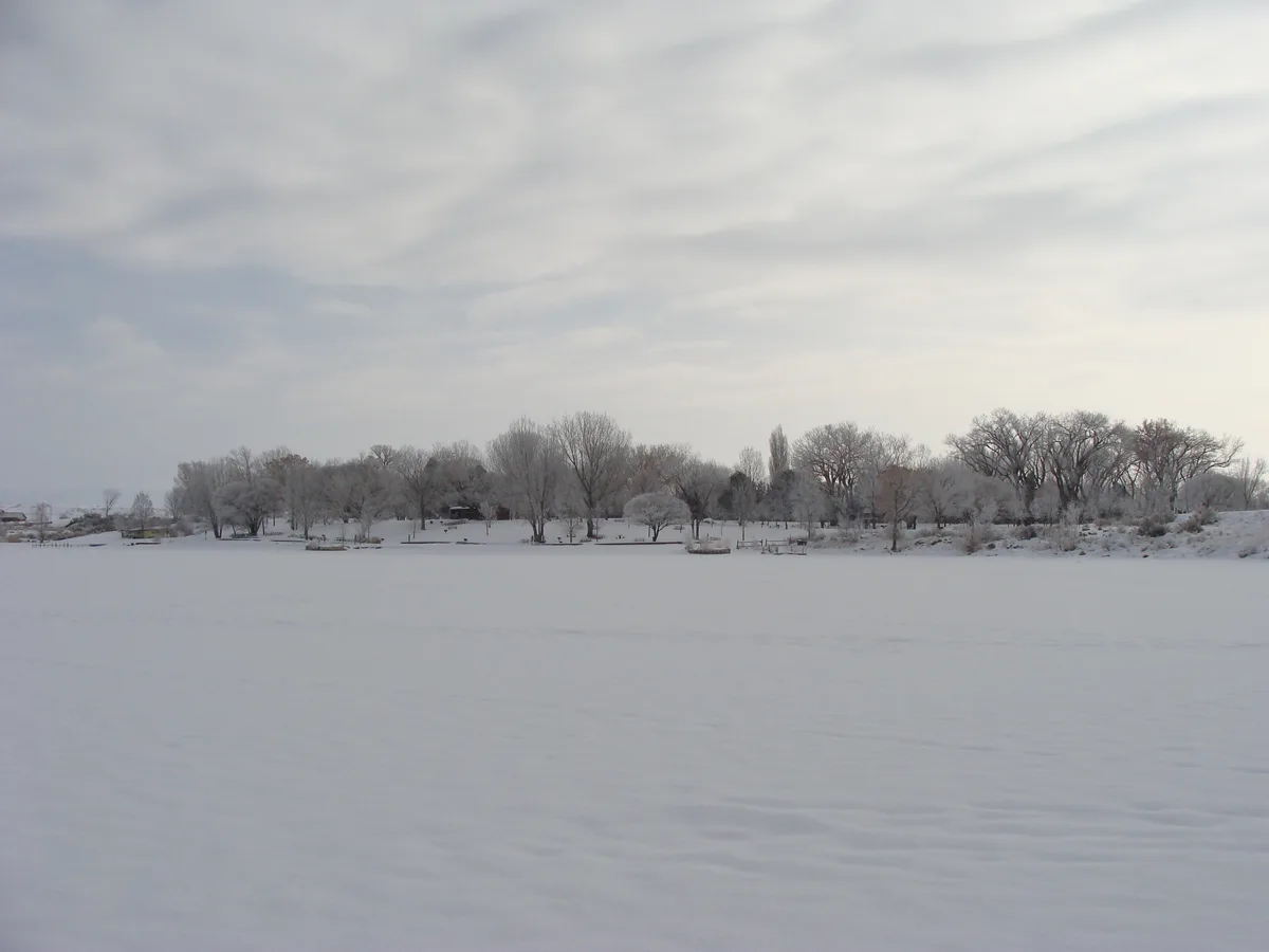 A wide winter view of Highline Lake shows a smooth, pale surface under a cloudy sky, with a line of frost-covered trees and shoreline structures across the far bank. The muted gray light gives the snowy landscape a quiet, still feel.