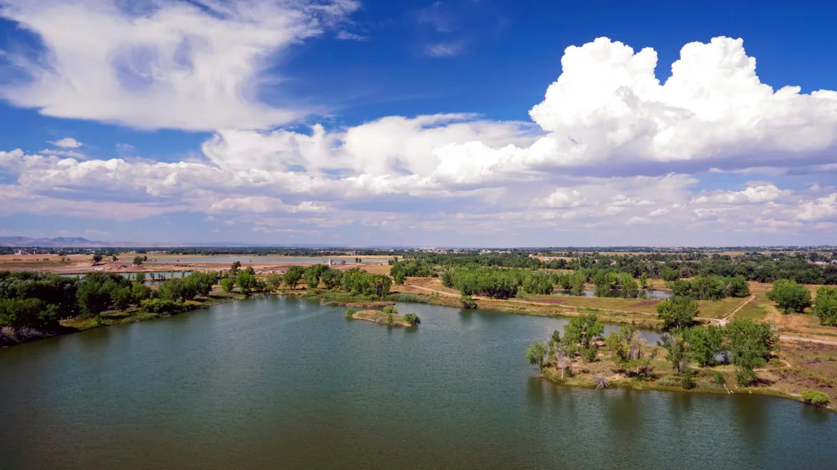 Wide view over a calm desert lake bordered by low green trees and sandy shoreline, with open water in the foreground and a flat, distant horizon under towering white clouds. The bright blue sky and still surface give the scene a spacious, reflective feel.