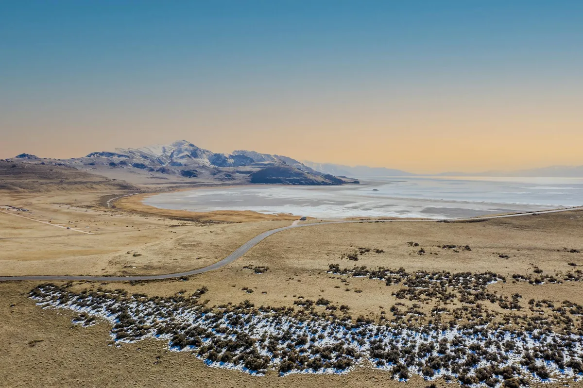 Wide view of a calm desert lake at golden hour, with pale water reflecting the surrounding low mesas and distant snow-dusted cliffs under a clear blue-to-gold sky. Sparse winter-brown grass and patches of snow line the shoreline, and a narrow road curves toward the water in the foreground.
