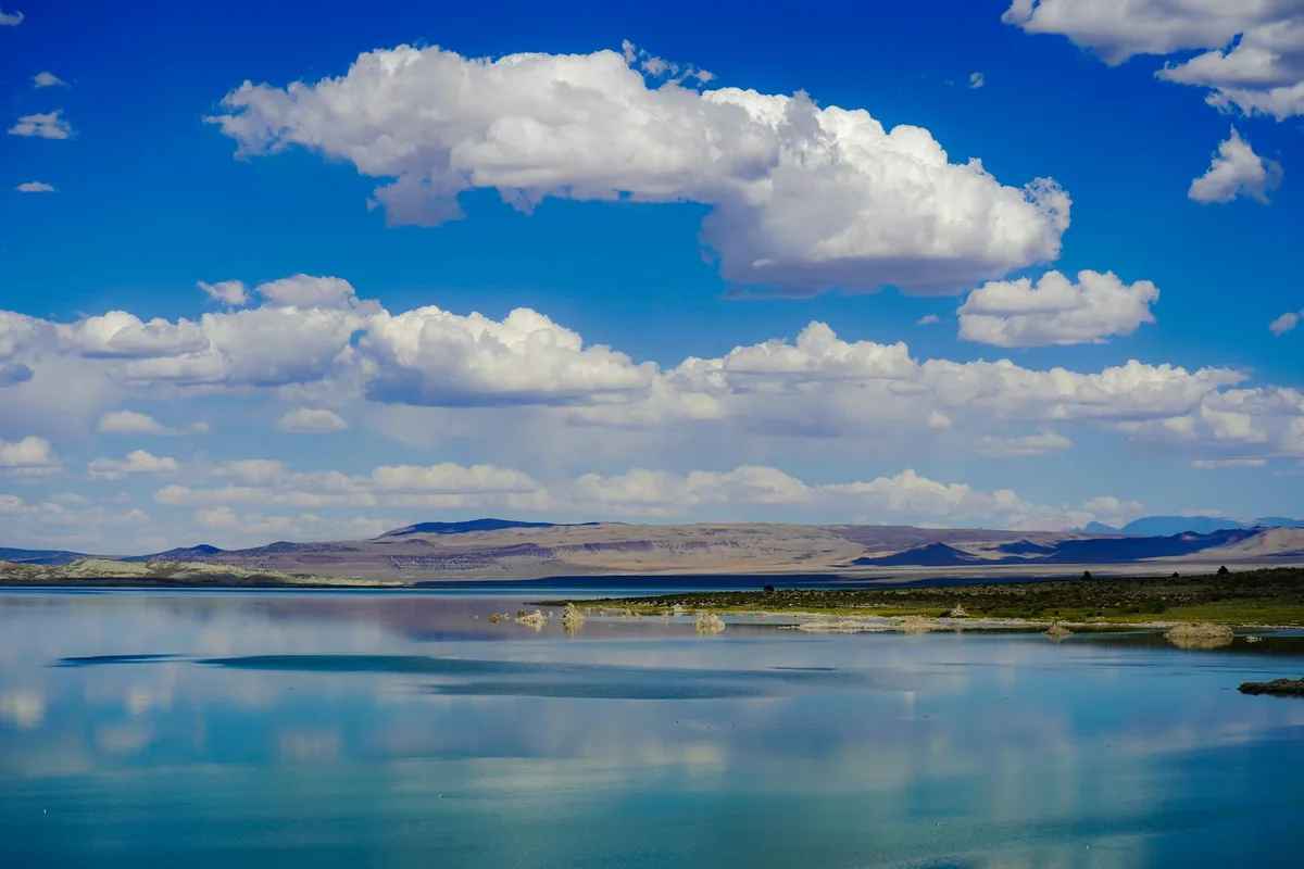 Calm blue water at Highline Lake reflects a wide sky filled with bright cottony clouds, with low desert mesas and cliffs stretching across the horizon. Sparse shoreline vegetation and a distant ridge line emphasize the park’s open, high-desert setting.