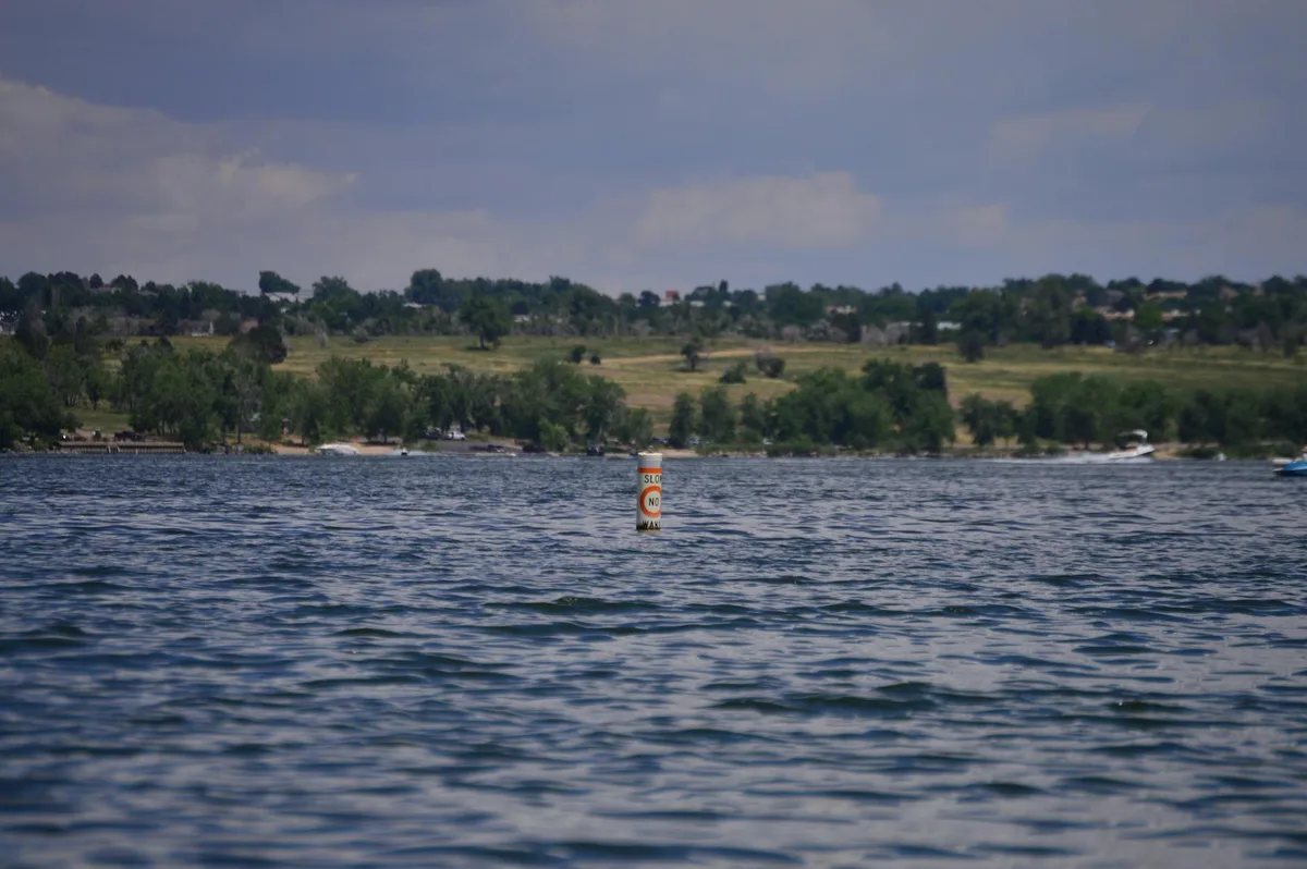 Low, rippled blue water fills the foreground, with a small buoy marked “Slow No Wake” standing near the center of the lake. Across the shore, a green, tree-lined hillside rises under a hazy sky with a few soft clouds.