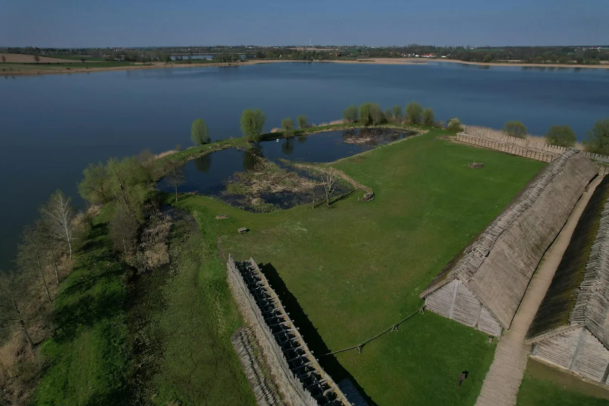 Wide aerial view of the Holzwarth Historic Site on a grassy riverbank, with weathered log buildings and a fenced corral beside small ponds and the broad blue Colorado River. The scene is bright and open, with spring-green trees and the valley stretching into the distance under a clear sky.
