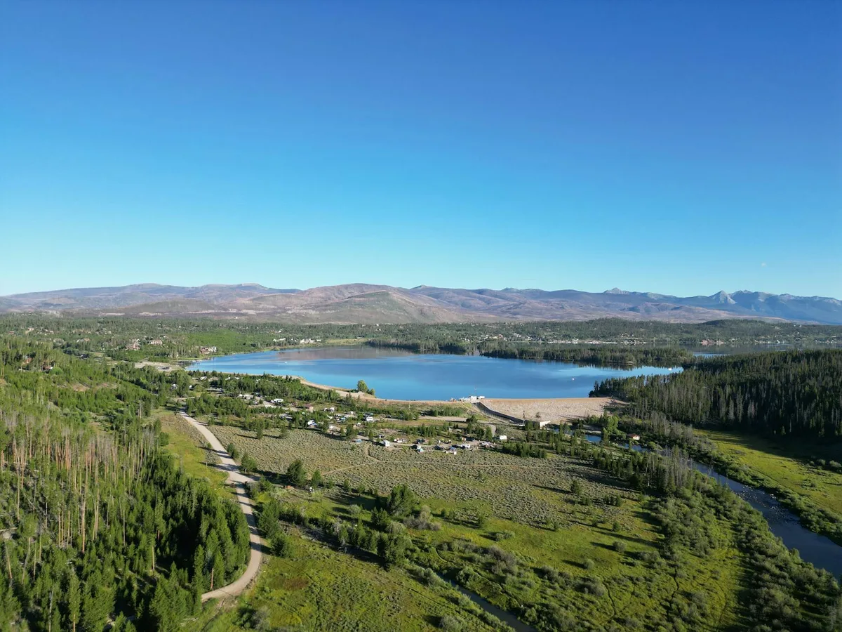 A sweeping aerial view shows Holzwarth Historic Site’s small cluster of rustic cabins spread across a green meadow beside the Colorado River, with a wide blue lake and forested shore in the middle distance. Low mountain ridges and the rugged Never Summer range stretch across the horizon under a clear, bright sky.