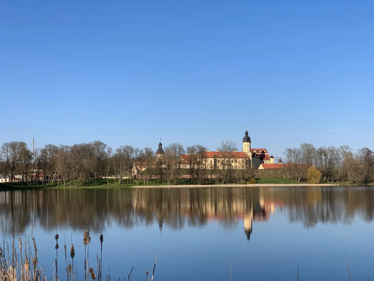 A calm lake mirrors a long historic complex with red roofs, pale yellow walls, and a tall tower behind a line of bare trees. Dry reeds frame the foreground, and the clear blue sky and still water give the scene a crisp, early-spring feel.