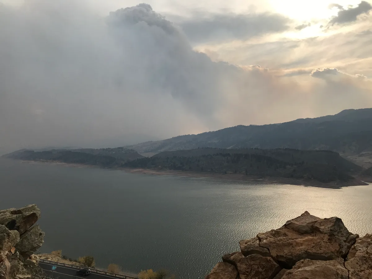 Golden-hour light glows on Horsetooth Reservoir as a thick plume of smoke rises over the foothills and hazy clouds fill the sky. Dark ridges and rocky foreground outcrops frame the calm water, which reflects a bright strip of sunlight near the shore.