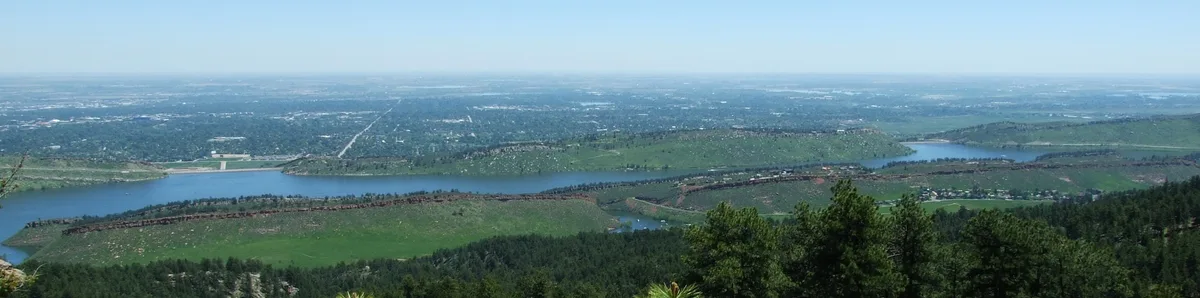 A wide panoramic view shows Horsetooth Reservoir stretching through green foothills and rocky hogbacks, with the water cutting across the landscape beneath a pale blue sky. The foreground is filled with dark evergreen trees, while the distant plains fade into a hazy horizon.