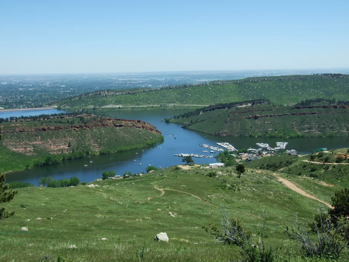 Wide view over Horsetooth Reservoir framed by rolling green foothills and red-rock ridges, with the inlet curving around a marina packed with white boats. Clear blue sky and sunlit slopes give the scene a bright, open feel.