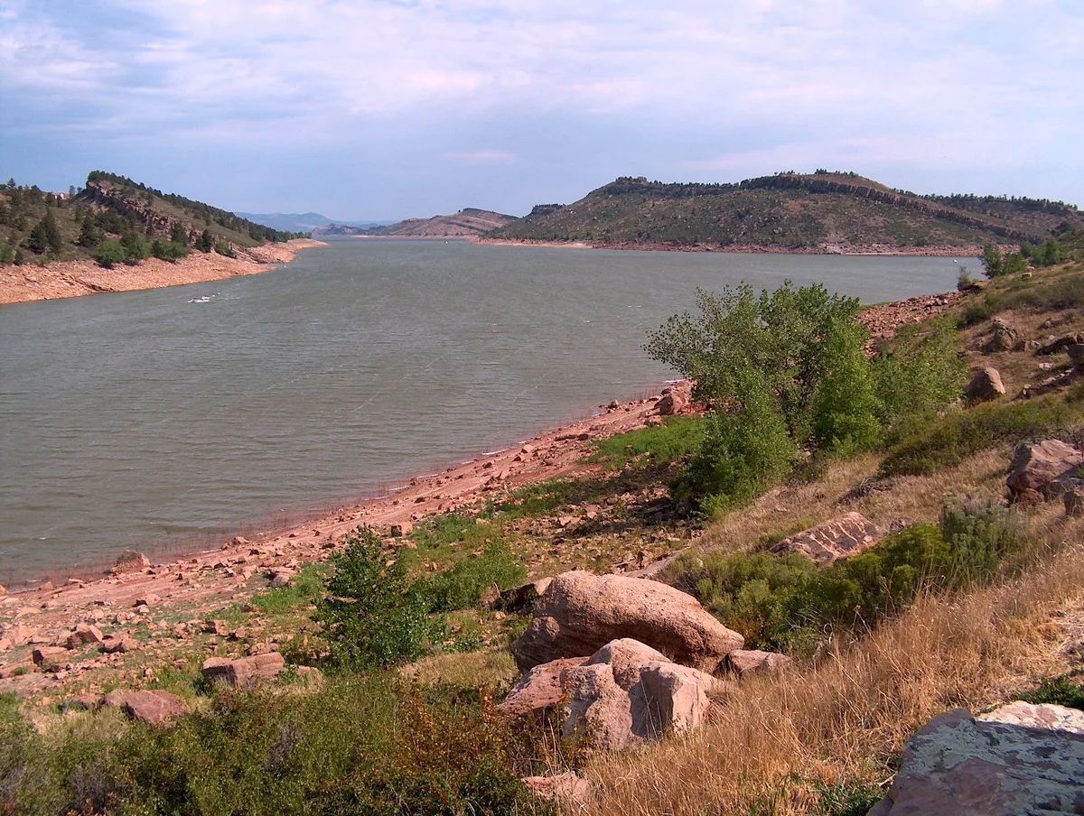 Wide view of Horsetooth Reservoir stretching between rocky, shrub-covered foothills, with pinkish shoreline stones in the foreground. Calm gray water leads into the distance under a soft golden-hour sky with thin pastel clouds.