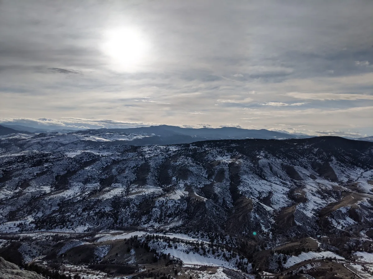 Snow-dusted foothills stretch across the frame under a bright, hazy sun and layered gray clouds, with dark ridgelines and winding roads cutting through the valley below. The wide view looks out over the winter landscape from high on Horsetooth Rock.