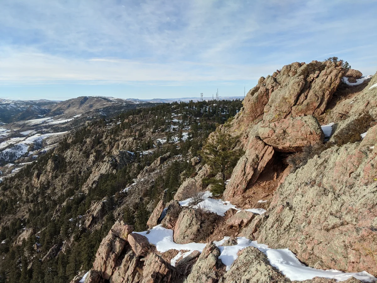Jagged Horsetooth Rock fills the right side of the frame, its pinkish granite ledges streaked with patches of snow and small evergreens clinging to the slopes. To the left, the view opens over snowy foothills and distant ridgelines beneath a pale blue sky with thin, wispy clouds.