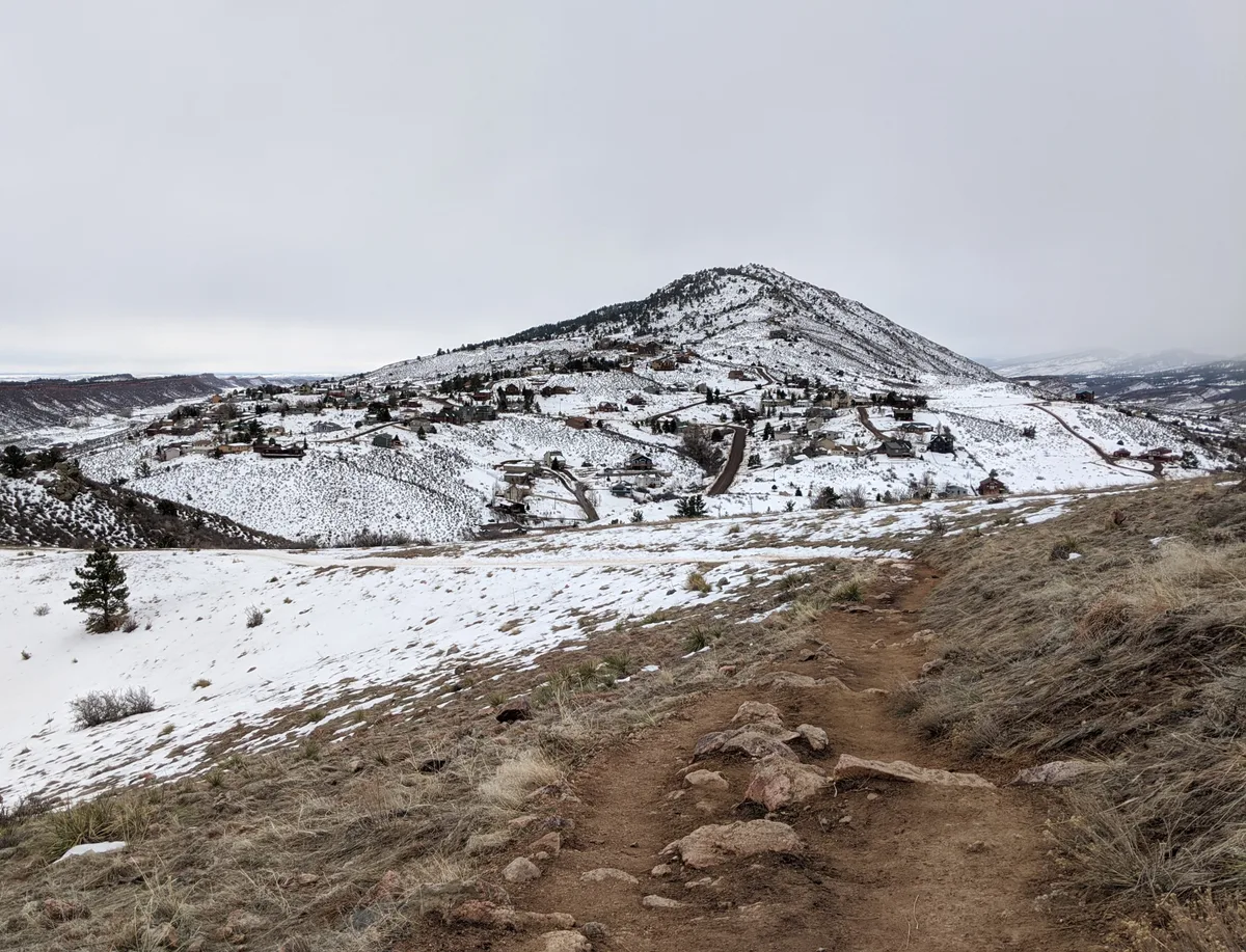 A rocky dirt trail leads down through patchy snow and brown winter grass toward the broad, snow-covered bulk of Horsetooth Rock, with scattered houses and roads spread across the foothills below. The overcast sky flattens the light, giving the scene a cold, muted winter look.