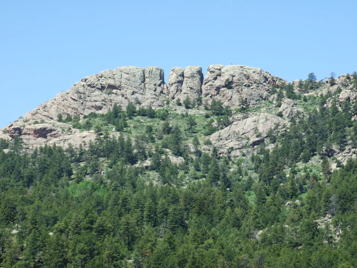 Horsetooth Rock rises above a green, pine-covered mountainside, its pale, blocky granite cliffs split by a narrow central notch against a clear blue sky. The wide view emphasizes the rugged foothills terrain and the formation’s distinctive horse-tooth shape.