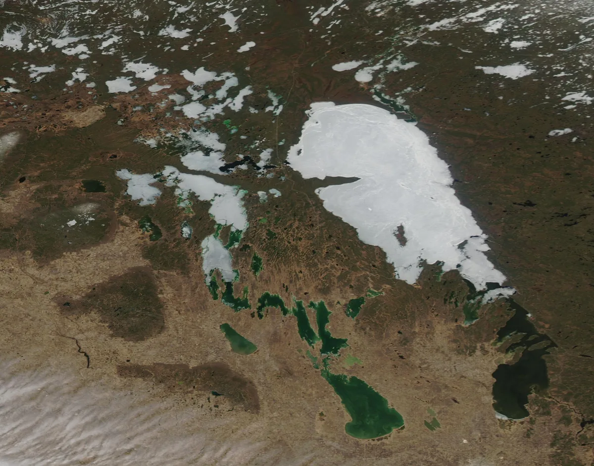 A high-altitude satellite view shows a large lake still partly covered in white ice, with smaller dark green lakes nearby set among brown, rugged terrain. Scattered snow patches and cloud shadow add texture across the mountain landscape.