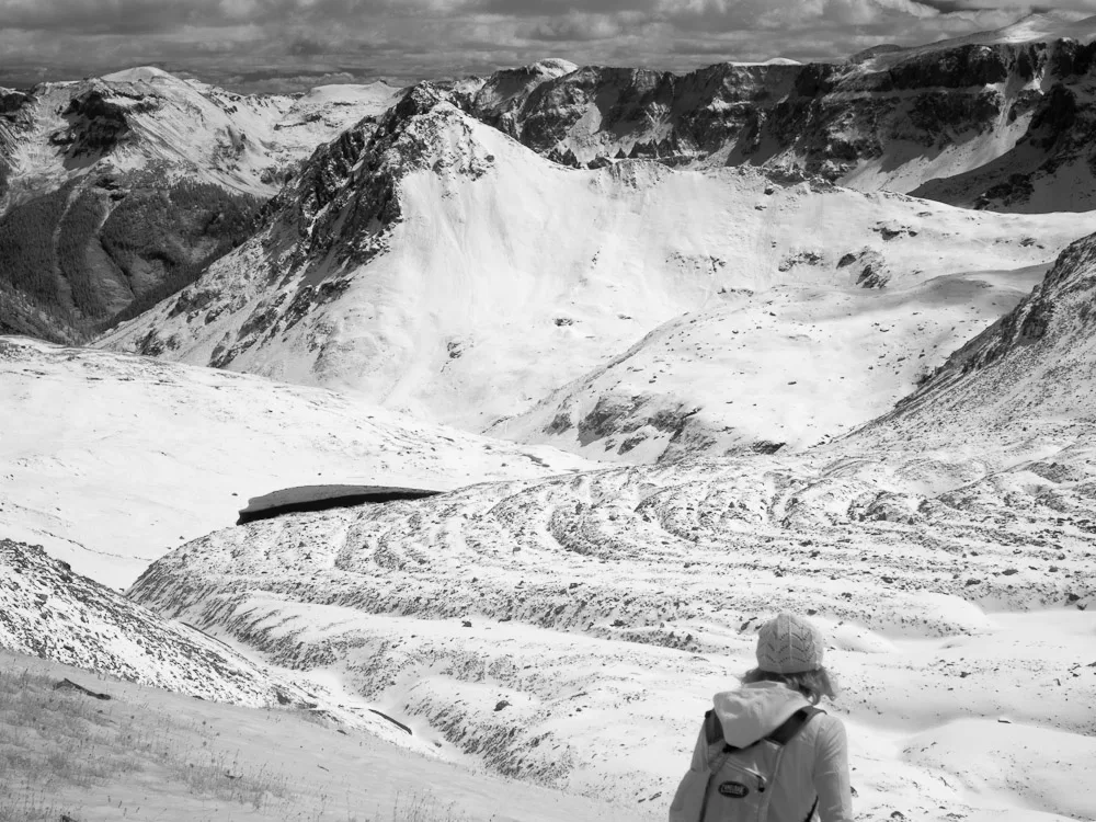 Wide view of a snow-filled alpine basin with steep, rugged peaks and dark rock ridges rising around a frozen or snow-covered lake. In the lower right, a lone hiker in a knit hat and backpack looks out over the bright winter landscape under a cloudy sky.