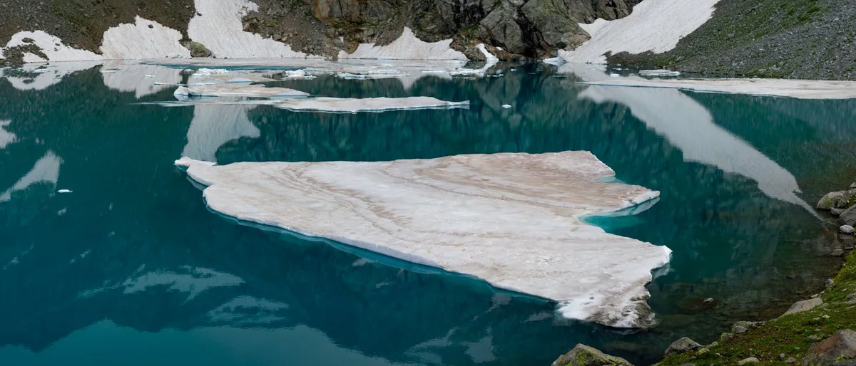 Turquoise alpine water fills a glacial basin, with a large white snow or ice floe floating in the foreground and smaller slabs scattered across the lake. Steep rocky slopes with lingering snow rise around the still surface, which reflects the dark cirque walls and sky.