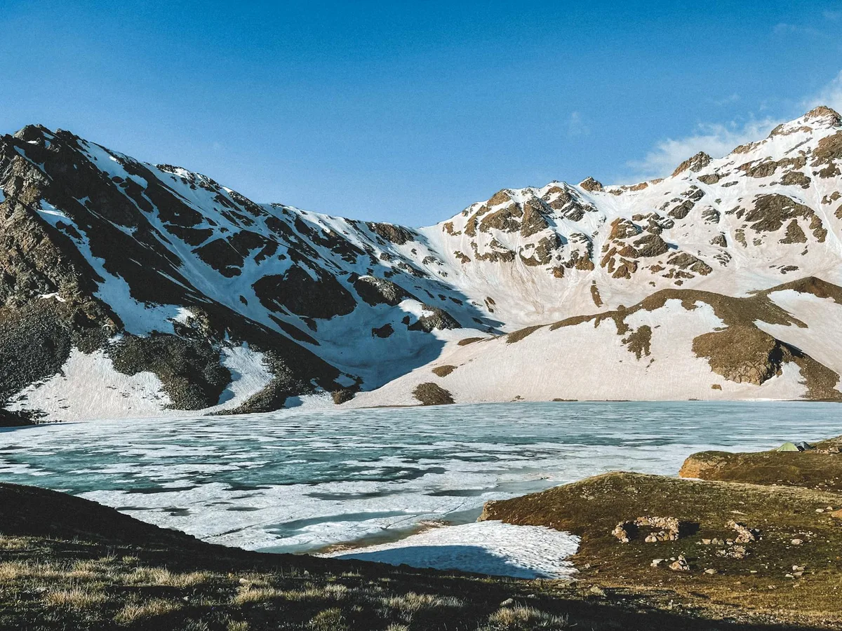 A partly frozen turquoise alpine lake sits beneath steep, snow-streaked cirque walls, with dark rock faces and white snowfields glowing in low golden light. Foreground patches of grassy meadow and exposed stone frame the icy water and rugged peaks.