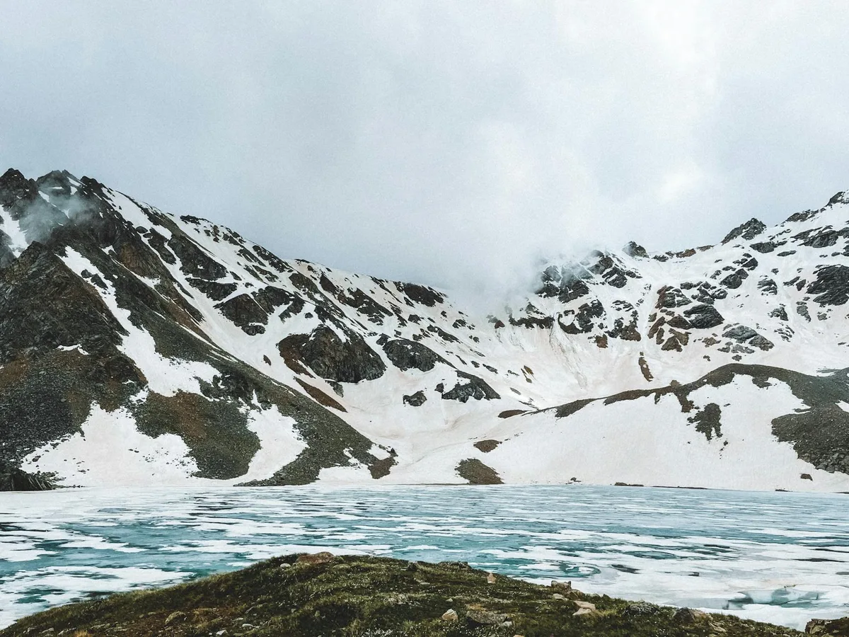A turquoise alpine lake with thin ice stretches across the foreground, backed by steep, snow-streaked mountain walls under a low cloudy sky. A small grassy ledge at the bottom edge adds a dark foreground contrast to the cold, glacial basin.