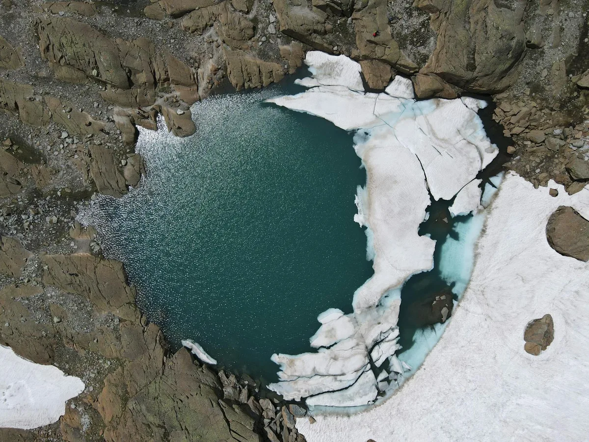 An overhead view of a turquoise alpine lake tucked into a rocky glacial cirque, edged by broad white snowfields and broken ice. Dark stone walls frame the water, with a narrow pale-blue meltwater channel cutting through the snow on the right.