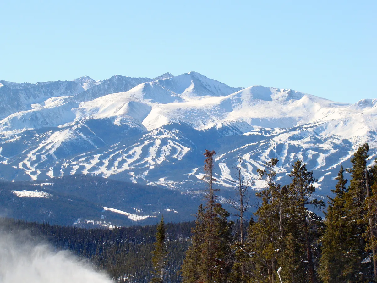 Snow-covered mountain peaks and ski runs fill the frame under a clear blue sky, with dark evergreen trees in the foreground. A low plume of mist or snow drifts up at the lower left, adding depth to the wide alpine view.