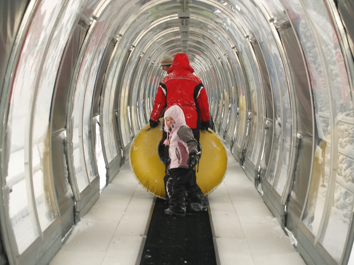 Two bundled-up skiers stand inside a clear, arched moving carpet tunnel at Keystone, with snow clinging to their jackets and boots. The transparent walls create a repeating reflections effect, and a bright yellow tube fills the center of the passage.