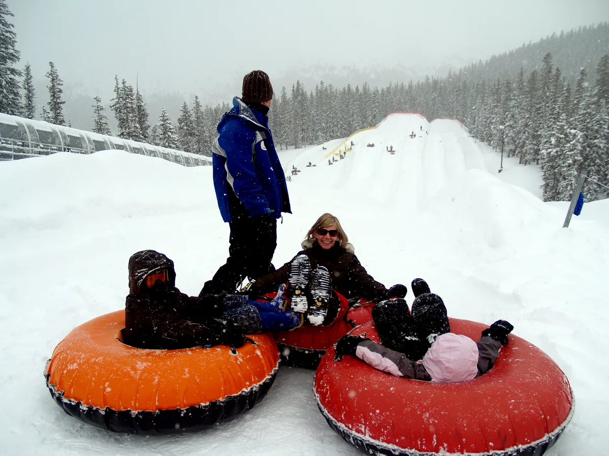 Family snow tubing on a snowy Keystone Resort slope, with three bright orange and red inner tubes clustered in the foreground and riders looking up the hill. Evergreen trees and a misty mountain backdrop frame the white tubing lanes under falling snow.