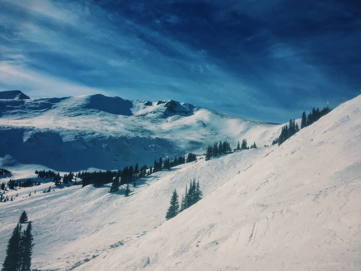 Snow-covered ski slopes rise toward rounded mountain peaks under a deep blue sky, with dark evergreen trees breaking up the white terrain. The wide view emphasizes the layered ridgelines and smooth, wind-shaped snow at blue hour.