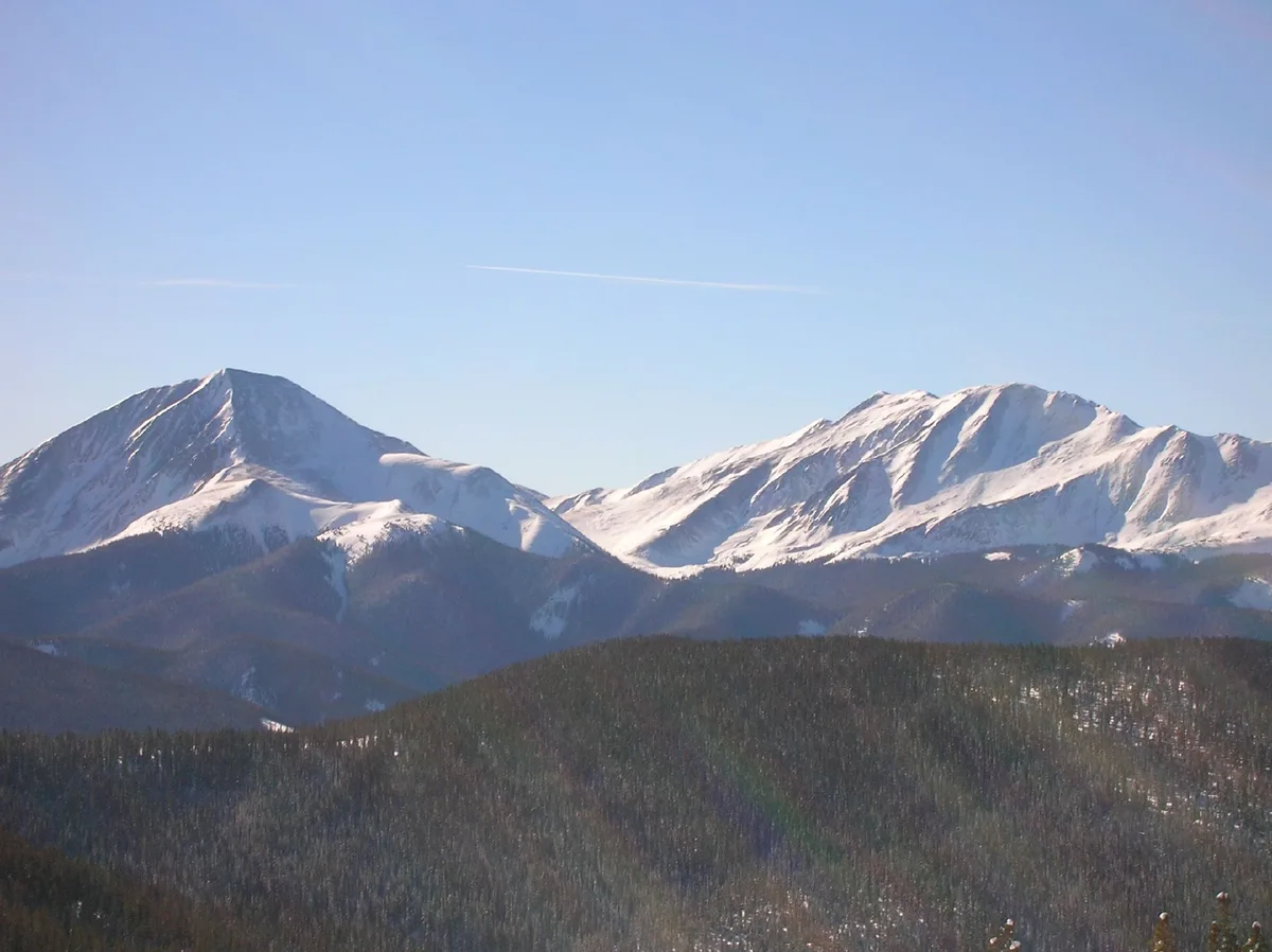 Snow-covered mountain peaks rise above dark forested ridges under a clear blue sky, with bright sunlight catching the snow on the upper slopes. The composition is a wide alpine landscape with no buildings or lake visible.