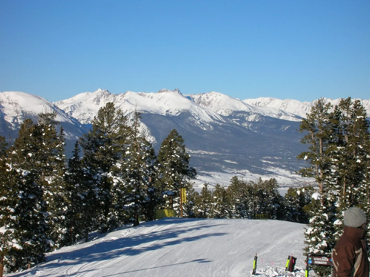 Snow-covered ski trail descending through pine trees with the Tenmile Range rising sharply in the background under a clear blue sky. A skier in a brown jacket stands at the right edge near a trail sign, with bright winter sunlight casting long shadows across the groomed snow.