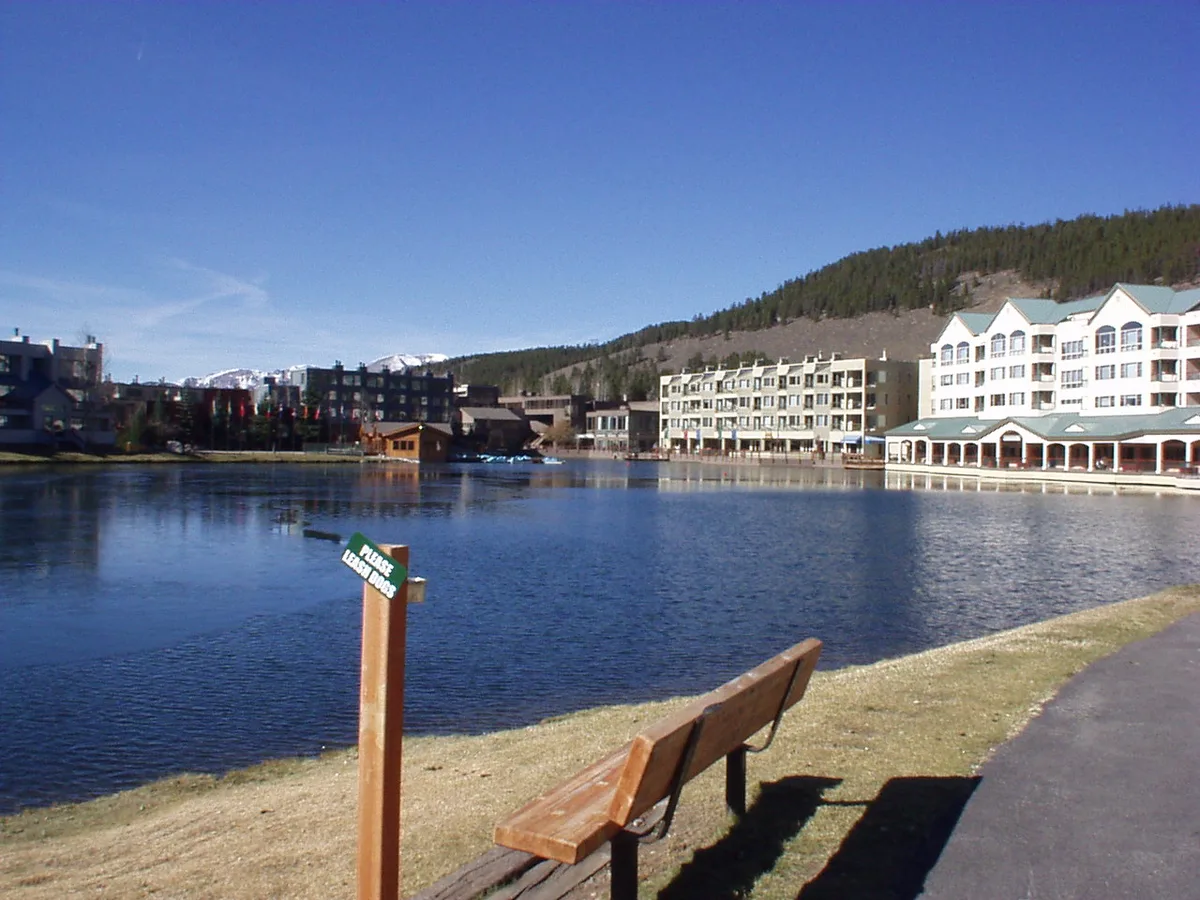 Blue-hour view across Keystone Lake with calm water reflecting the resort buildings and mountain slope beyond. A wooden bench and a small sign in the foreground line the paved path beside the lakeshore.