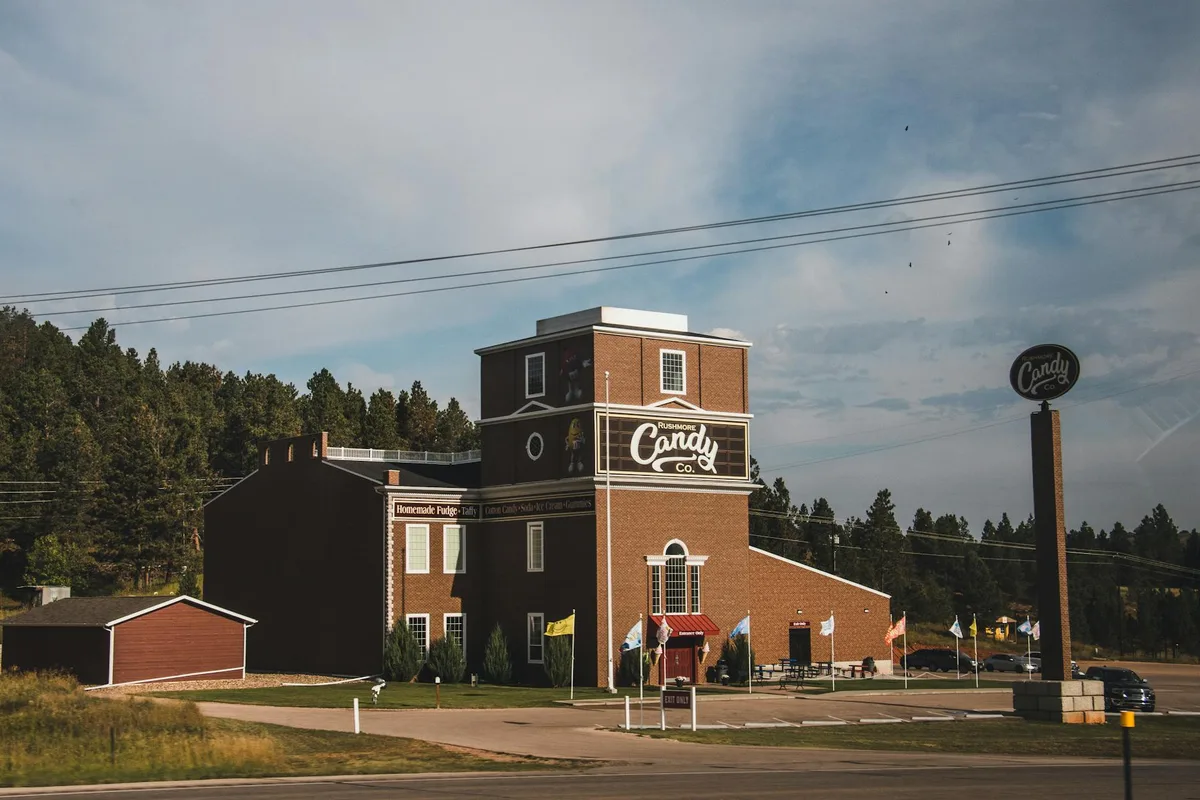 A rustic brick candy shop and adjacent dark-sided buildings sit beside a roadside lot, with a tall round “Candy Co.” sign, small flags, and pine-covered hills behind them. Soft blue-hour light and thin clouds give the scene a quiet, roadside mountain-town feel.