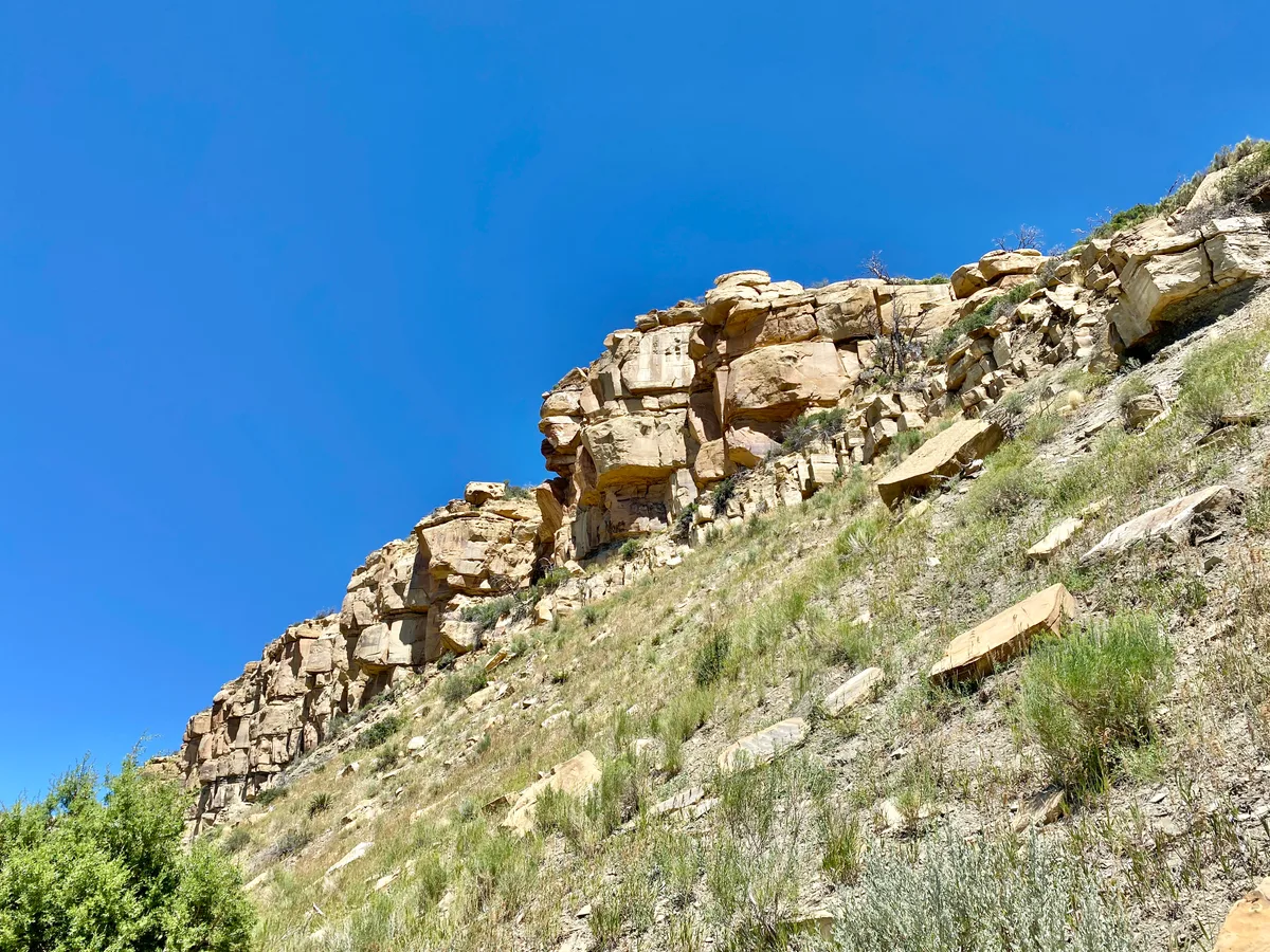 Sheer tan sandstone cliffs rise above a steep, grassy slope dotted with sage and scattered rock slabs, set against a clear deep blue sky. The composition emphasizes the rugged mesa edge and dry desert plateau textures.