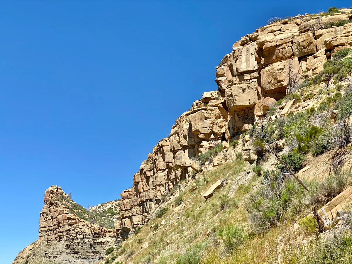 Sheer tan sandstone cliffs rise above a steep, scrub-covered mesa slope under a clear deep blue sky. A smaller rock outcrop stands in the distance, emphasizing the narrow, rugged edge of the road overlook.