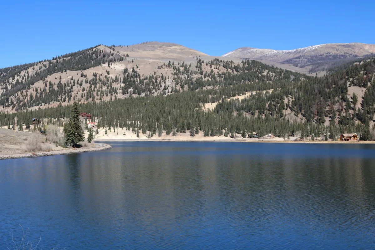 Wide view across Lake San Cristobal shows calm blue water in the foreground, with pine-covered slopes and pale, rounded mountains rising behind it under a clear blue sky. A few lakeside cabins and the shoreline curve along the far edge, with their shapes faintly reflected in the water.
