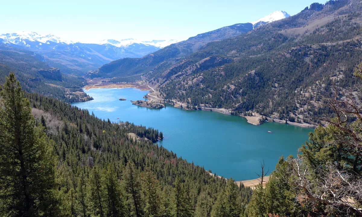 A wide overlook shows Lake San Cristobal’s bright blue water stretching through a steep mountain valley, bordered by dense evergreen forests and rocky slopes. Snowy peaks rise in the distance under clear light, with the lake’s calm surface reflecting the surrounding hillsides.