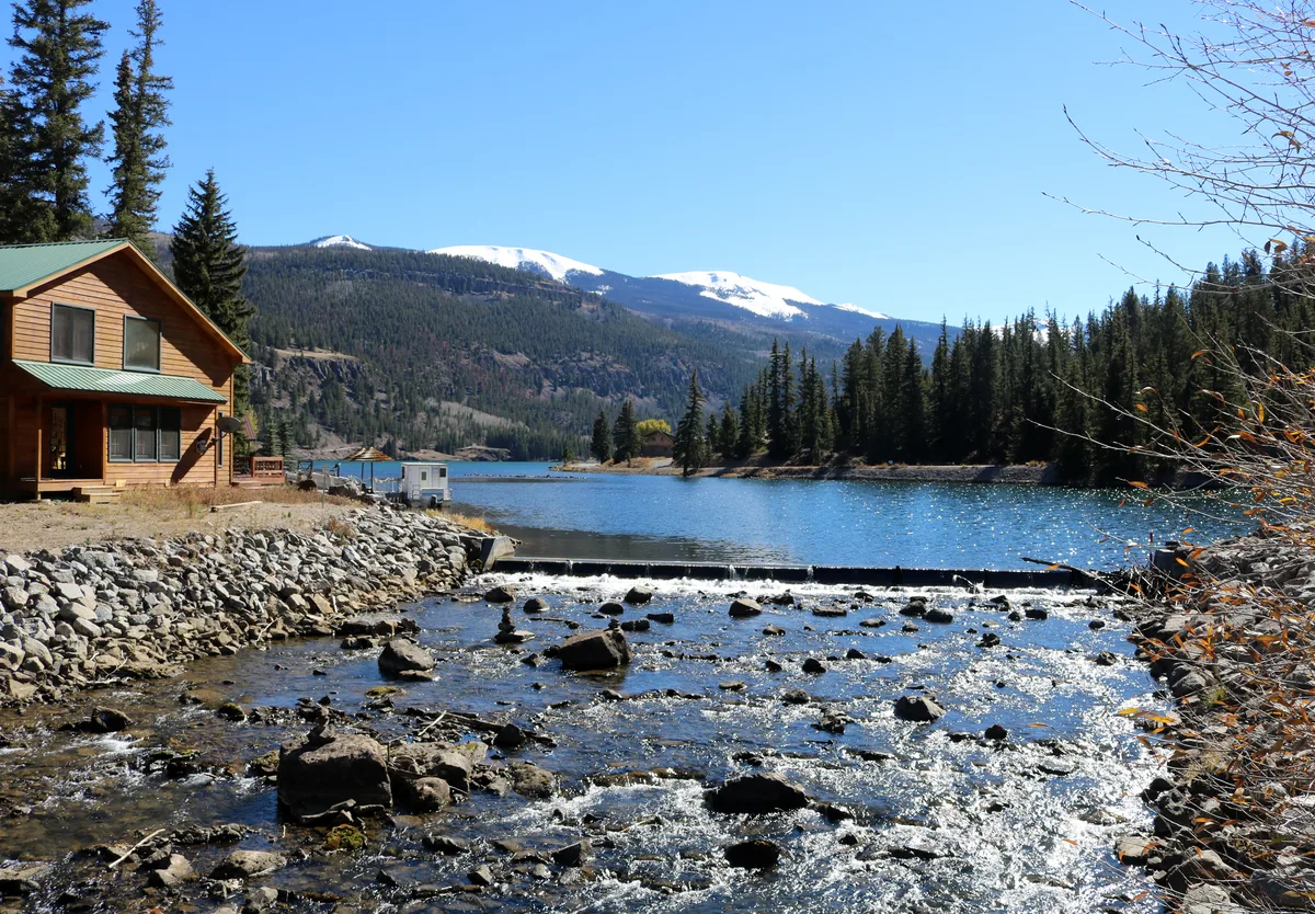 A wide view of Lake San Cristobal shows a rocky outflow stream in the foreground, with water sparkling in bright sunlight as it spills over a low dam. A cabin sits at the left edge beside the lake, while dark pine forests and snow-capped mountains rise across the blue water under a clear sky.