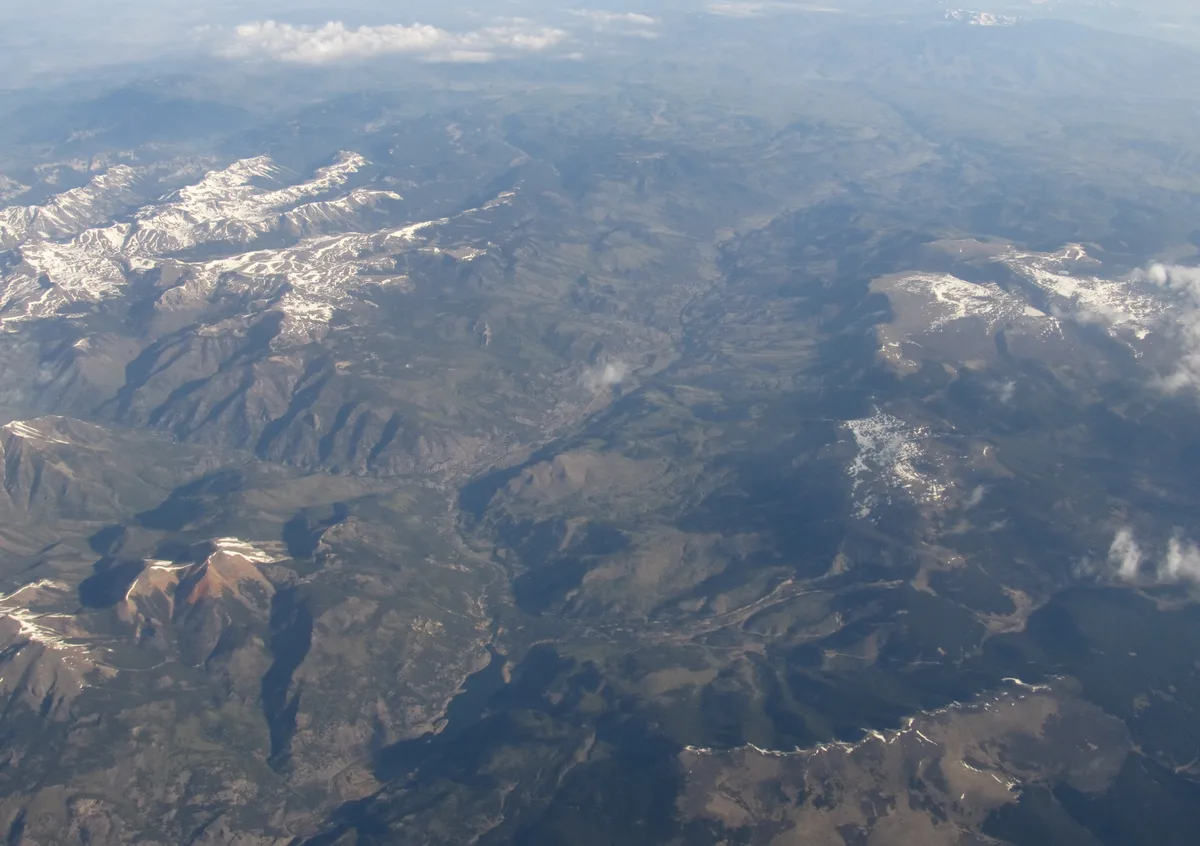 High above the mountains, snow patches cling to rugged ridges and dark valleys, with soft clouds and hazy light washing across the landscape. The wide aerial view emphasizes the layered terrain and sweeping scale of the peaks below.