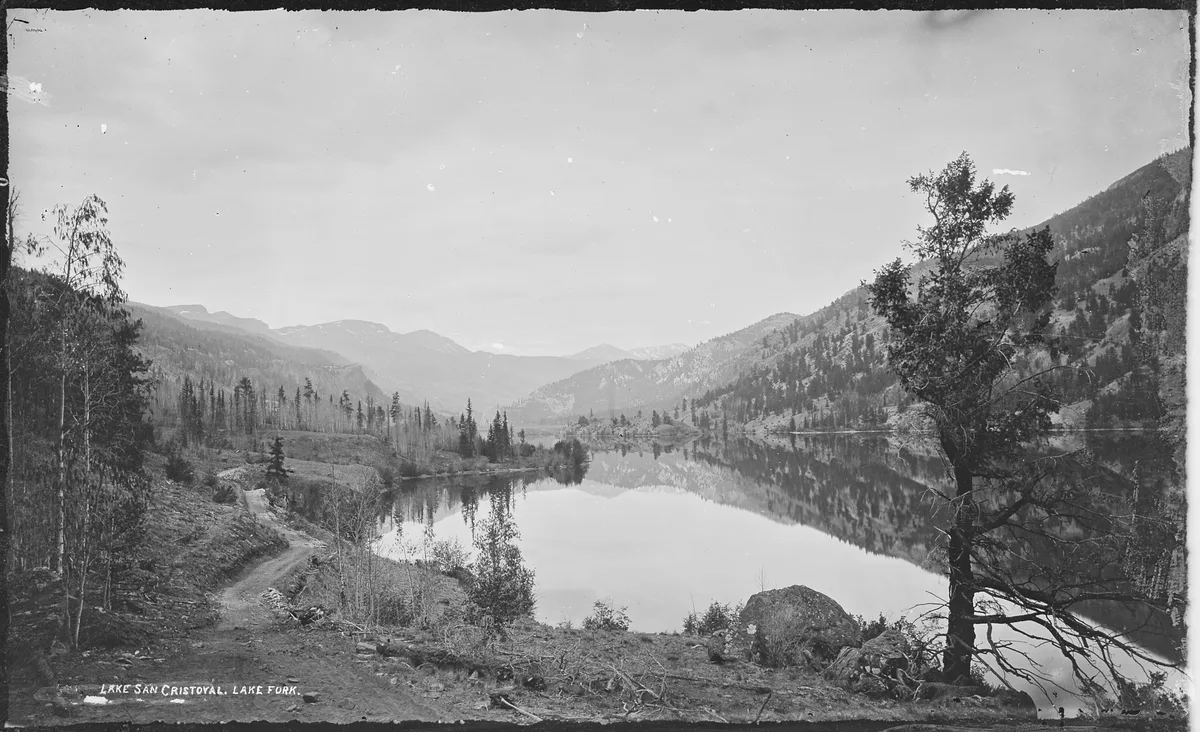 A wide view of Lake San Cristobal shows still water reflecting the surrounding forested mountains, with a narrow dirt path in the foreground and a lone tree and boulder along the rocky shore. The scene is quiet and expansive, with layered peaks fading into the distance under a bright, hazy sky.